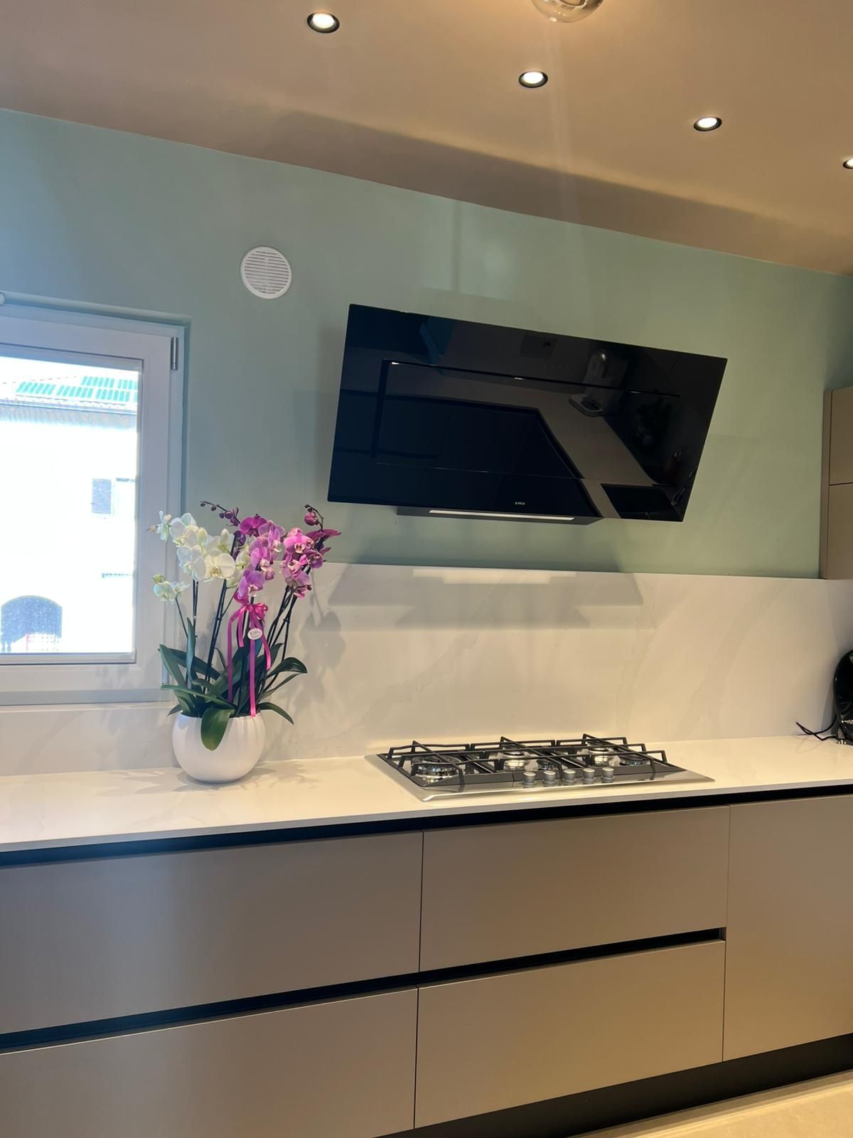 Cucina moderna con ripiani bianchi, mobili neri, TV a parete, piano cottura e fiori rosa vicino a una finestra.