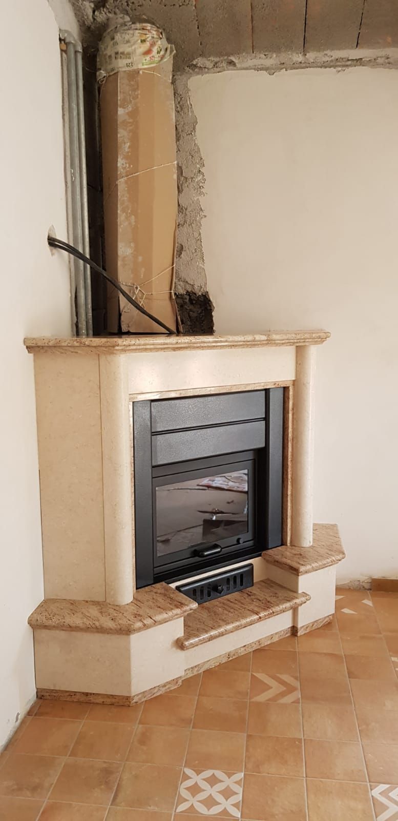 Camino beige con inserto scuro in una stanza piastrellata, con canna fumaria che si erge alle sue spalle.