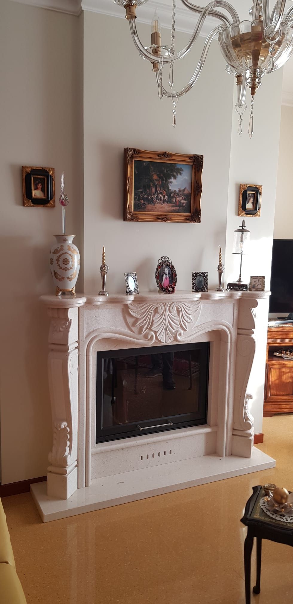 Elegante soggiorno con camino bianco decorato, lampadario e quadri appesi alle pareti.