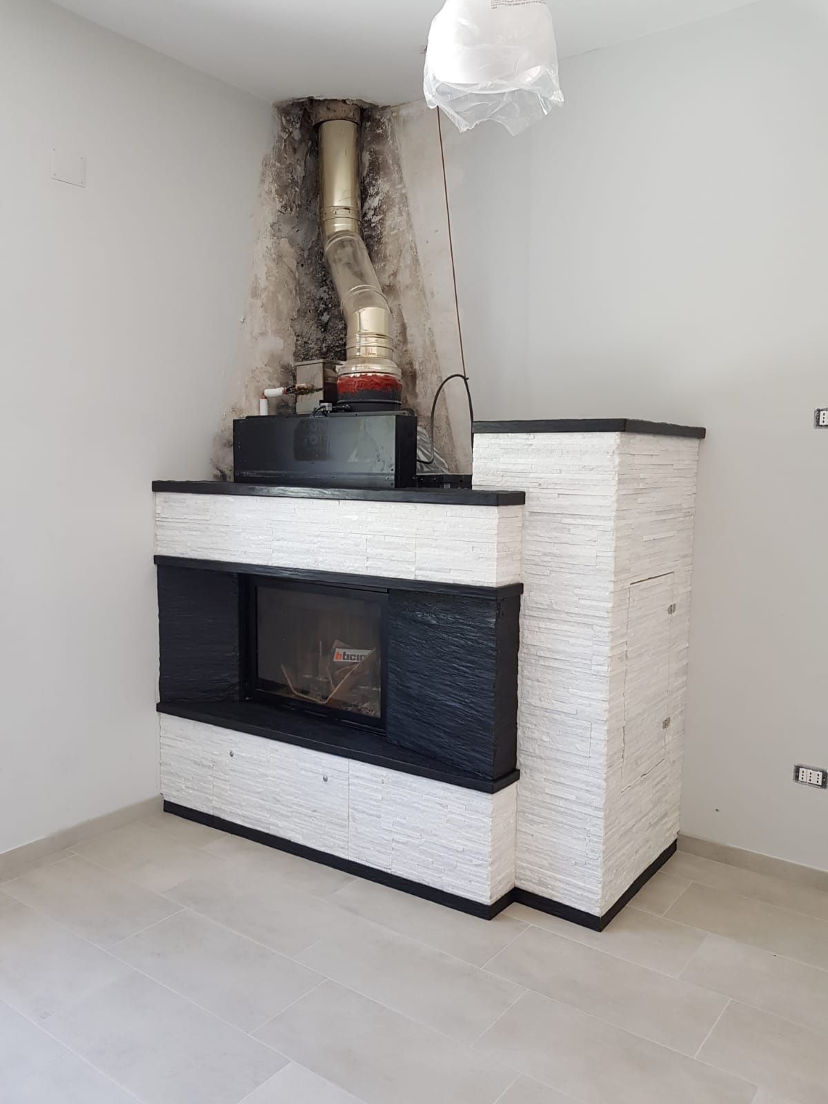 Caminetto moderno bianco con finiture nere e tubi di ventilazione a vista in una stanza vuota.