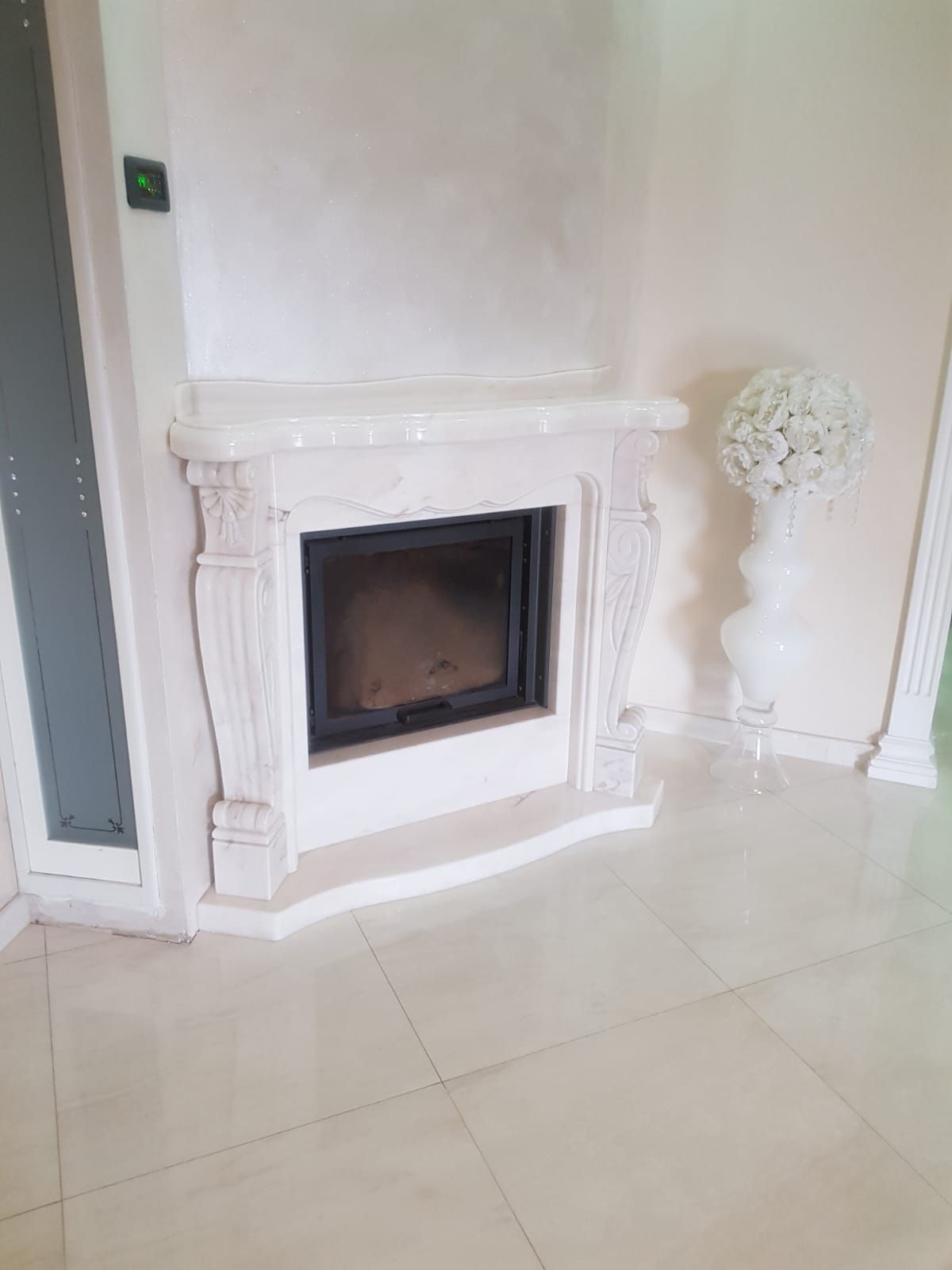 Caminetto decorativo bianco con focolare scuro in una stanza luminosa con pavimento piastrellato lucido.