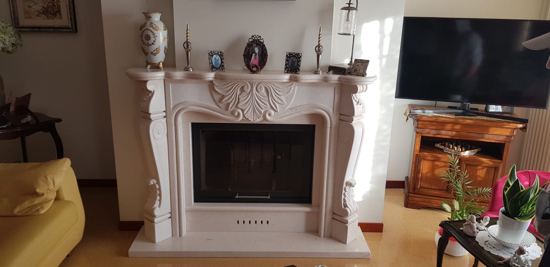 Soggiorno con camino bianco decorato, televisore su supporto in legno, divano beige e fiori su un tavolino da caffè.