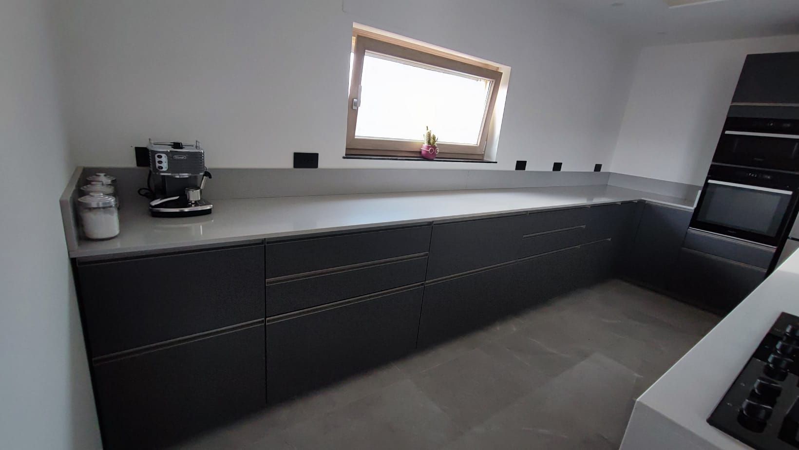 Cucina moderna con lunghi pensili neri, piano di lavoro bianco e una piccola finestra sopra il lavello.