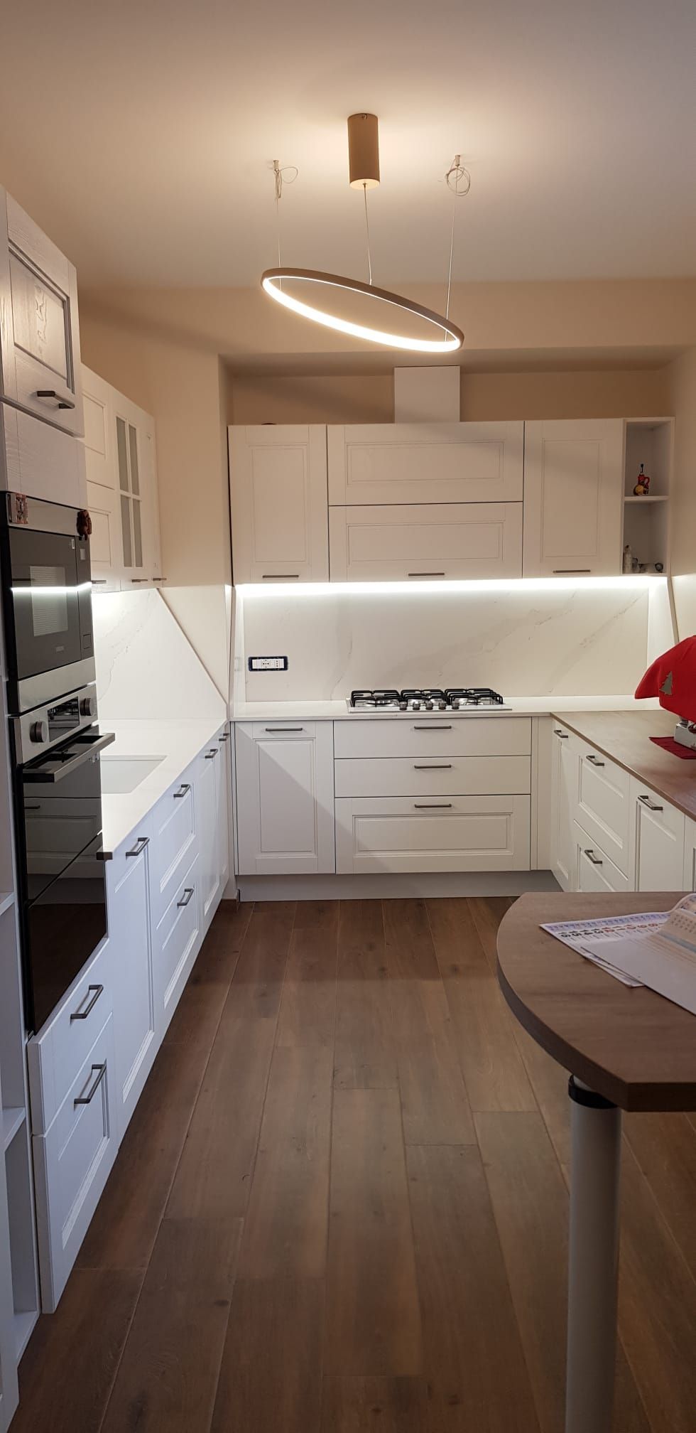 Cucina moderna bianca con pavimento in legno, armadi a muro, isola e lampada a sospensione.