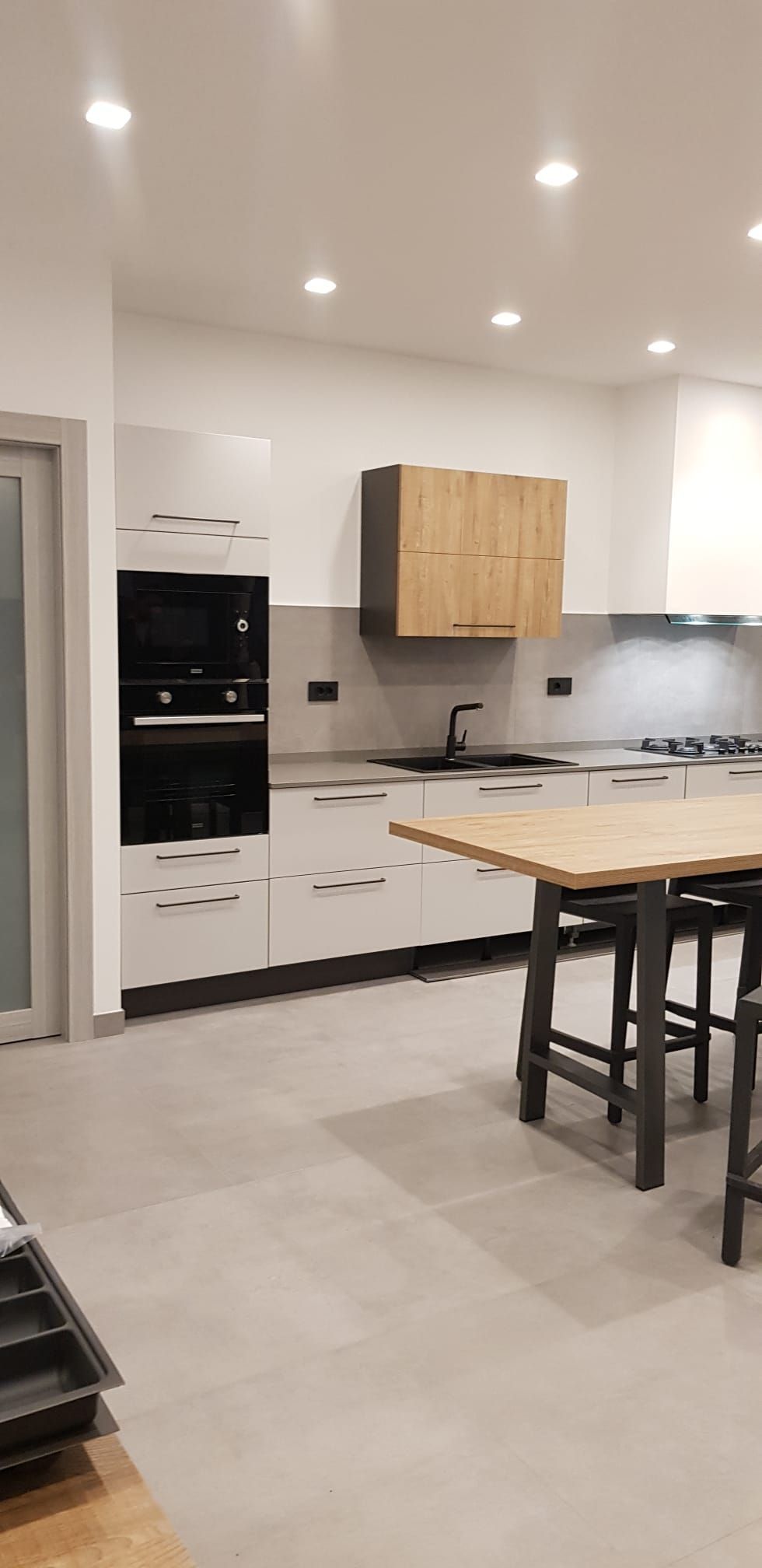 Cucina moderna bianca con isola, mobili in legno chiaro, elettrodomestici a incasso e faretti a soffitto.