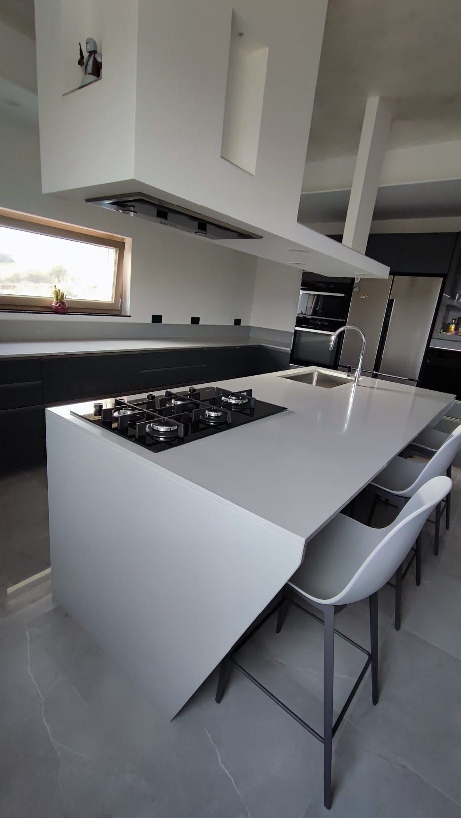 Cucina moderna bianca con isola, piano cottura a gas, sgabelli da bar e ampia finestra vicino al lavello.