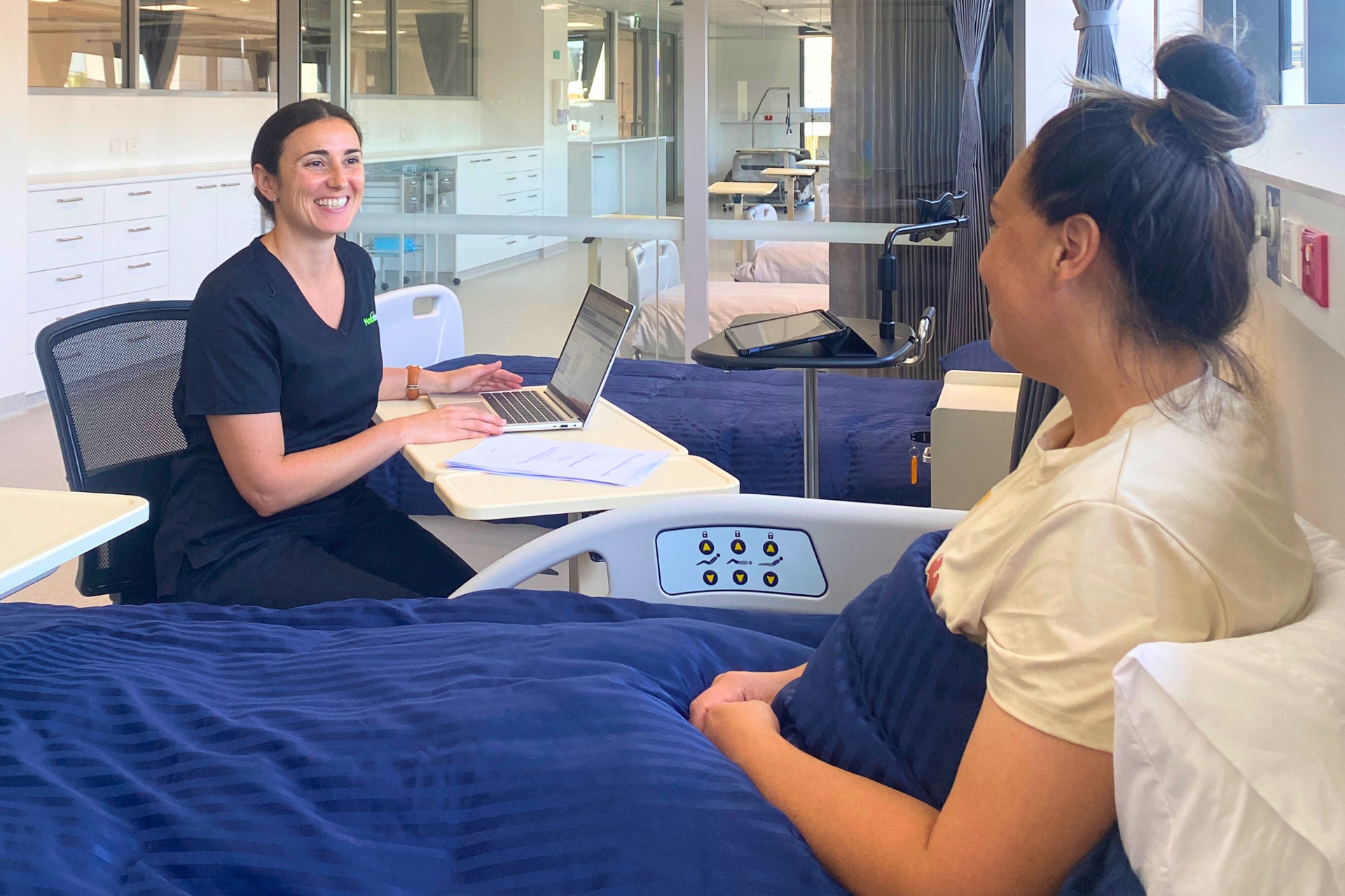 Woman in scrubs with a laptop smiles at a patient in a hospital bed.