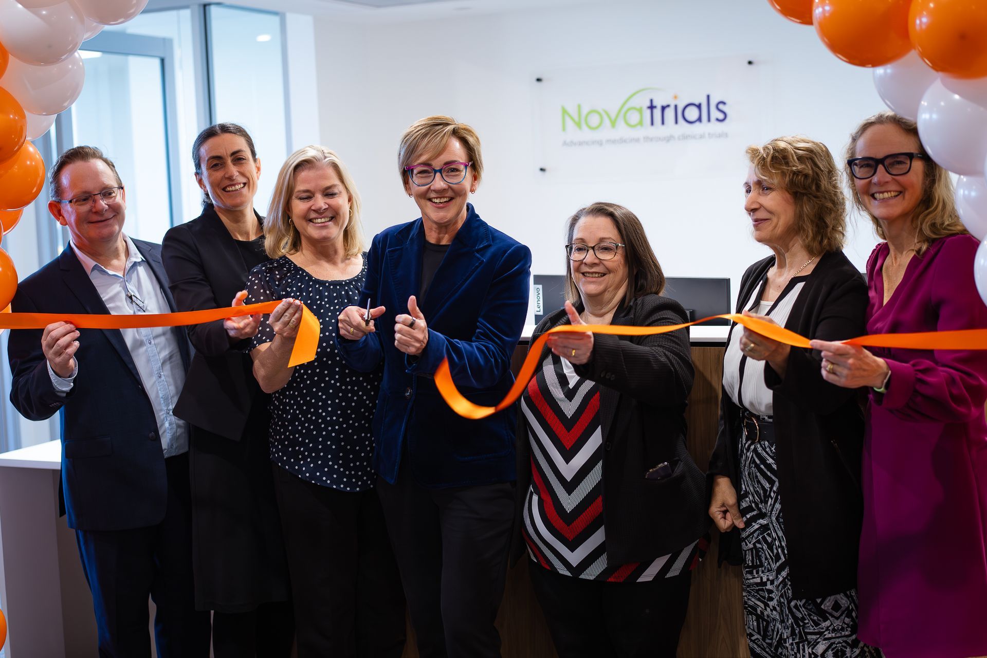 Ribbon-cutting ceremony with six people holding an orange ribbon in front of a Novatrials sign