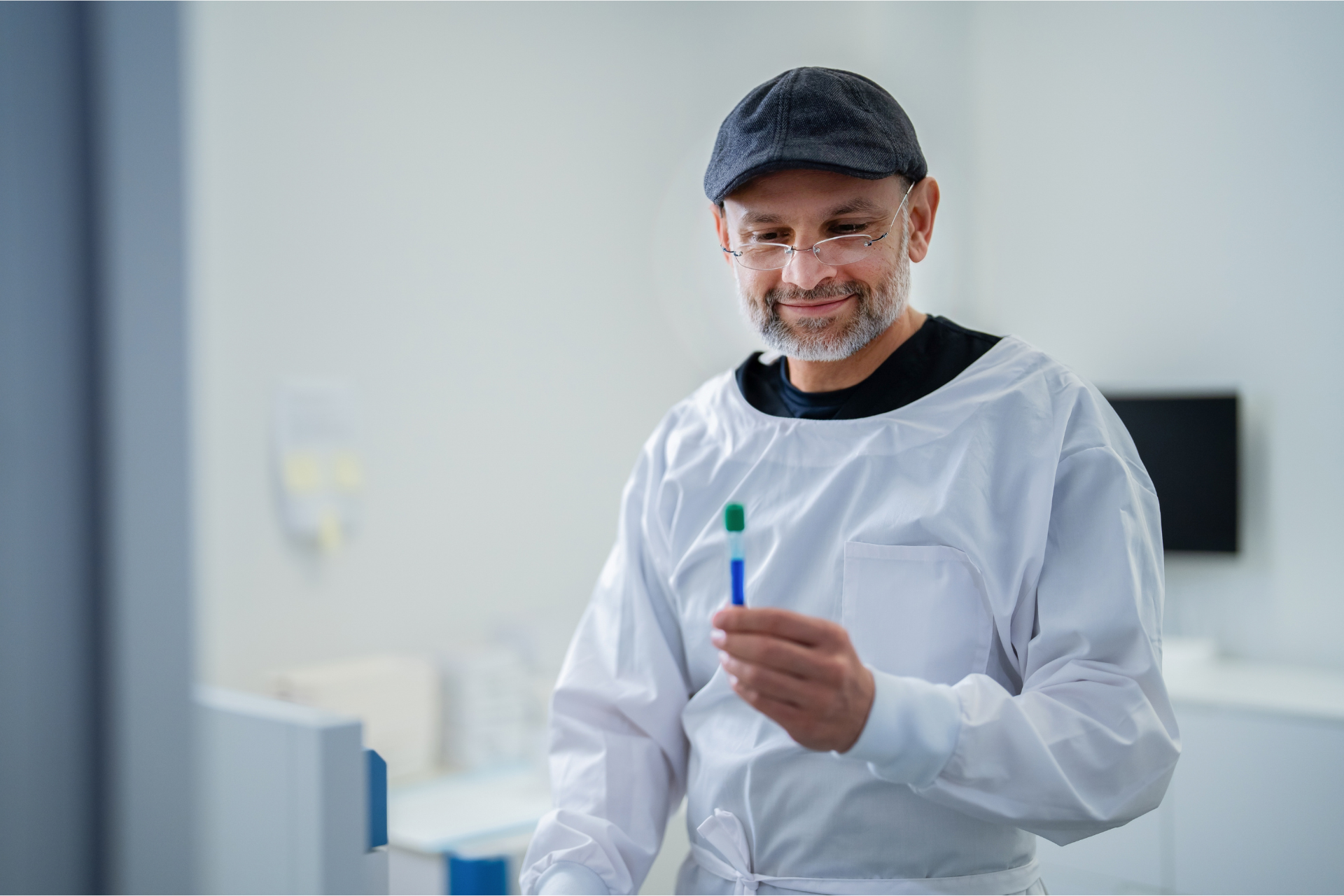 Man in lab coat, holding a test tube, smiling. Wearing a cap and glasses, in a medical setting.