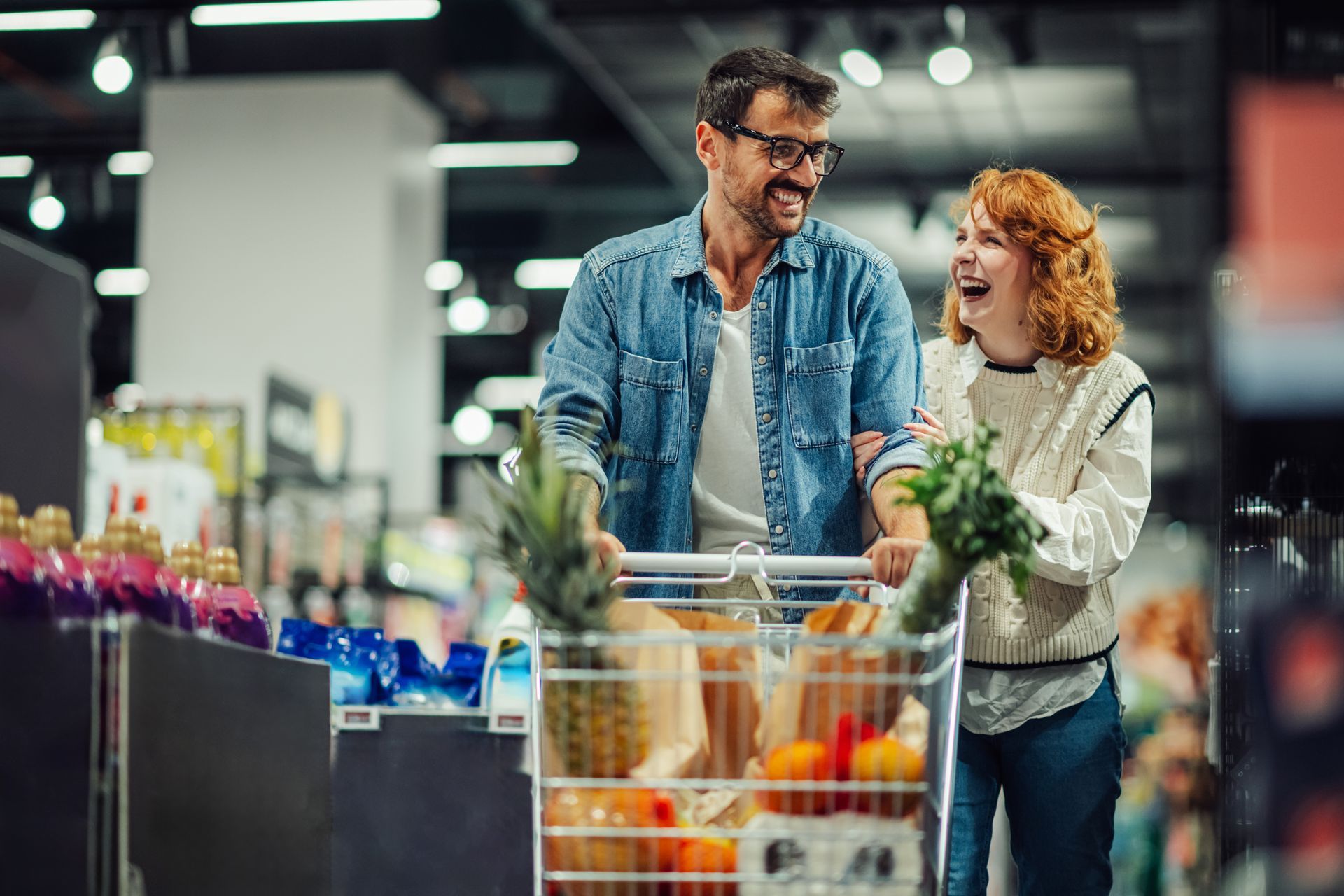 Two shoppers smiling over a grocery cart in a supermarket, with fresh produce and flowers nearby