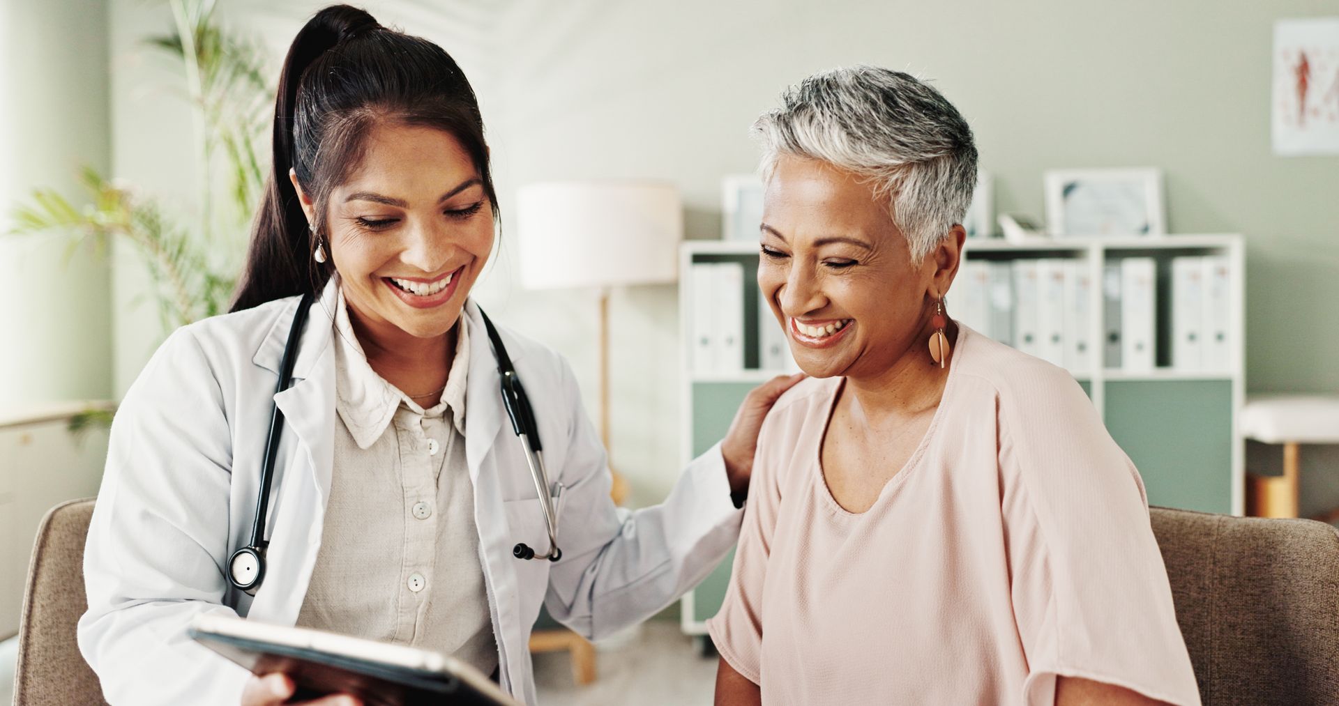 Doctor smiling and showing a tablet to a seated patient in a bright clinic office