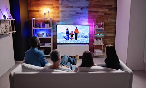 People on a couch watching a TV in a cozy living room with a lit lamp and wood-paneled wall