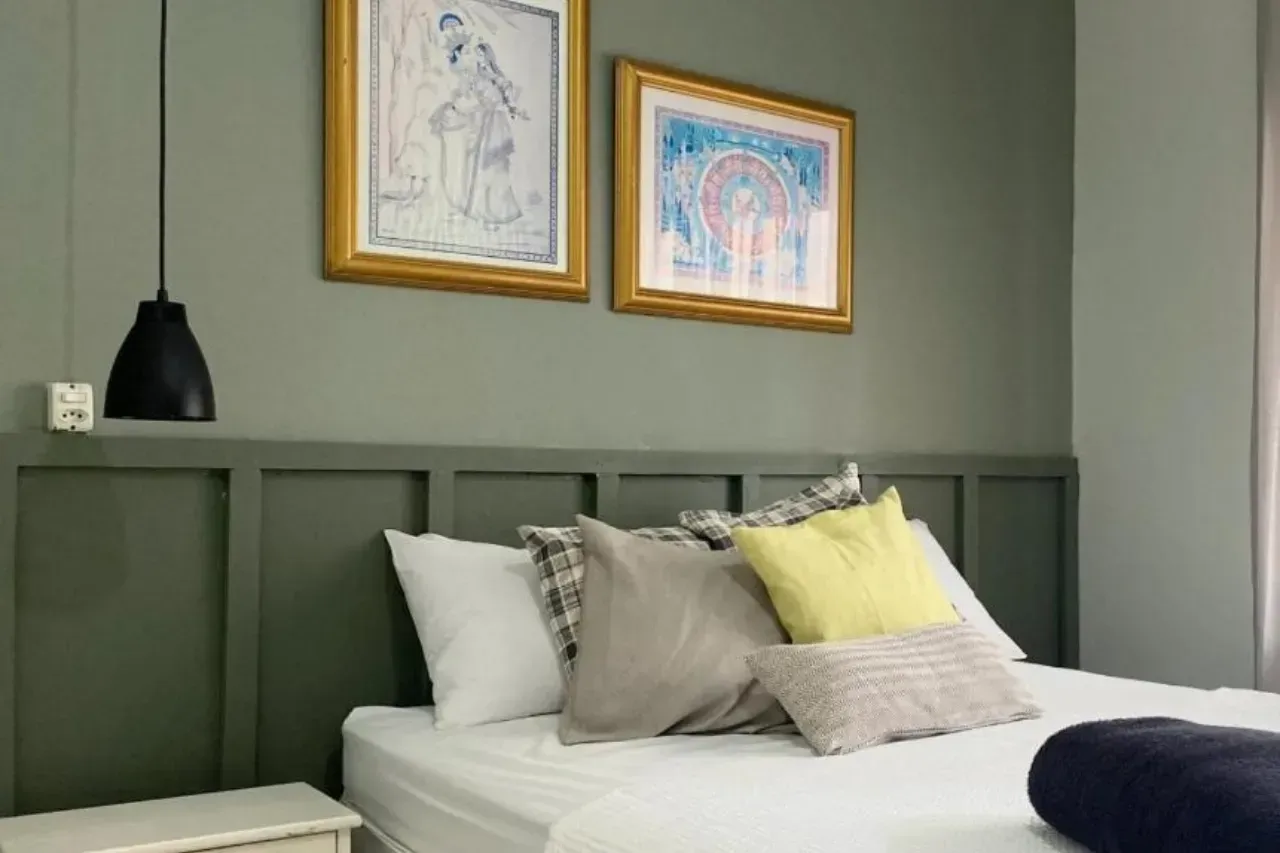 Cama arrumada num quarto verde suave com obras de arte emolduradas e um candeeiro de cabeceira preto.