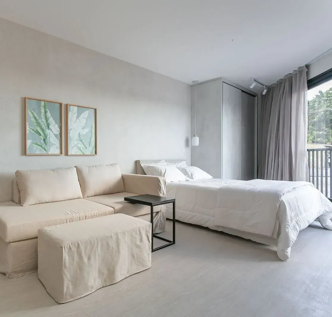 Quarto minimalista e luminoso com sofá creme, cama, mesinha de centro e cortinas cinza junto à porta da varanda.