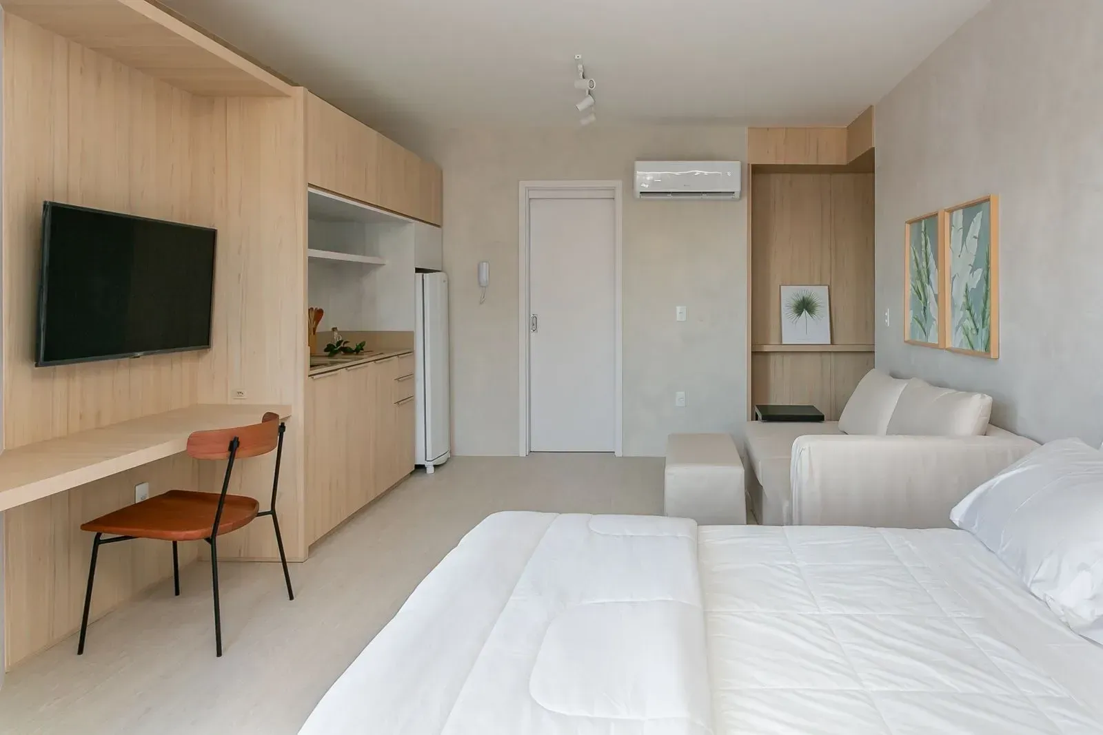 Quarto de hotel moderno com duas camas, TV de parede, sofá, escrivaninha e armários de madeira clara.