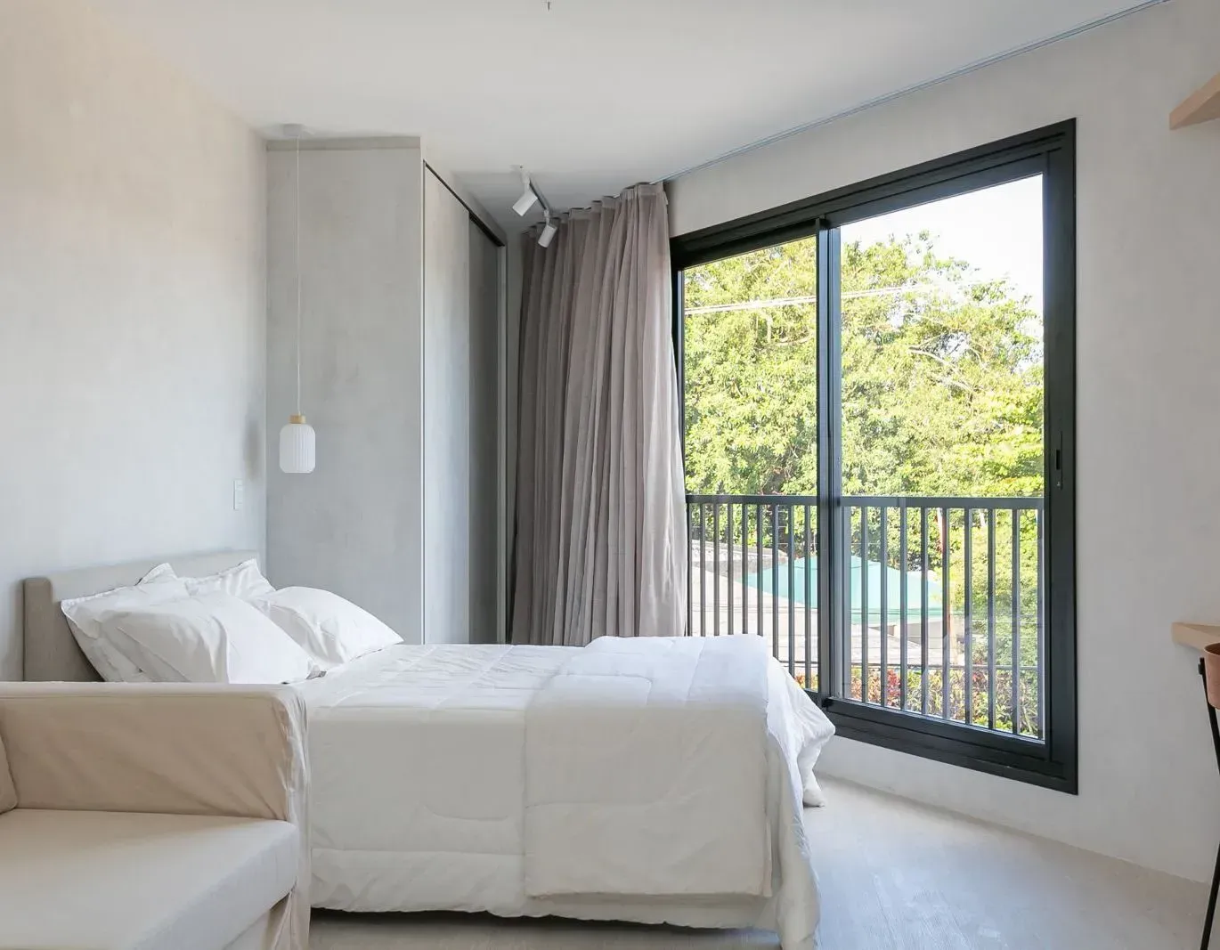 Quarto minimalista com cama branca, cortinas cinza e janela ampla com vista para a vegetação.