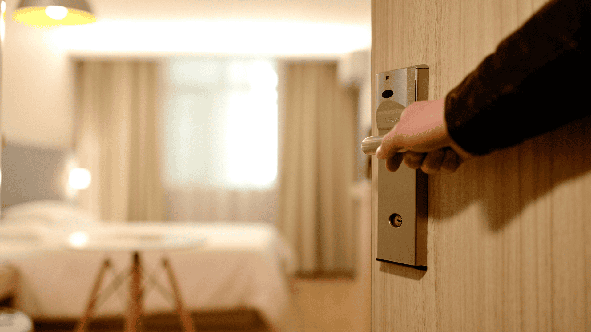 Mão abrindo a porta de um quarto de hotel, com uma cama suavemente iluminada e cortinas ao fundo.