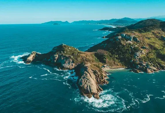 Península costeira rochosa com ondas turquesas do oceano e ilhas distantes sob um céu límpido.