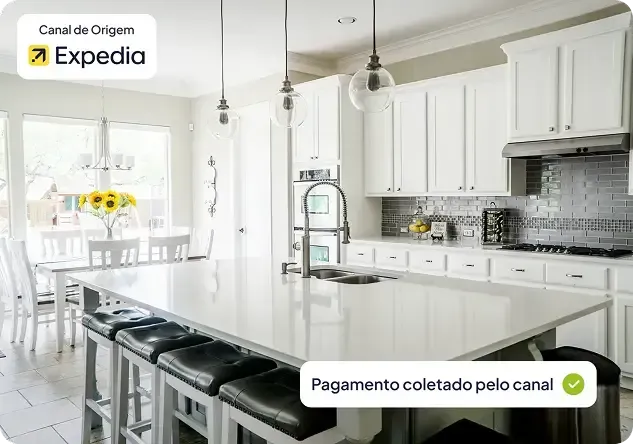 Cozinha branca e iluminada com ilha central, luminárias pendentes e área para refeições junto à janela.