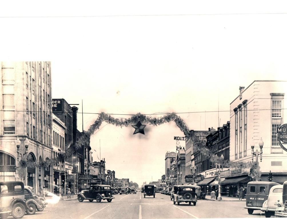 Historic black-and-white city street with vintage cars and buildings decorated with garlands.