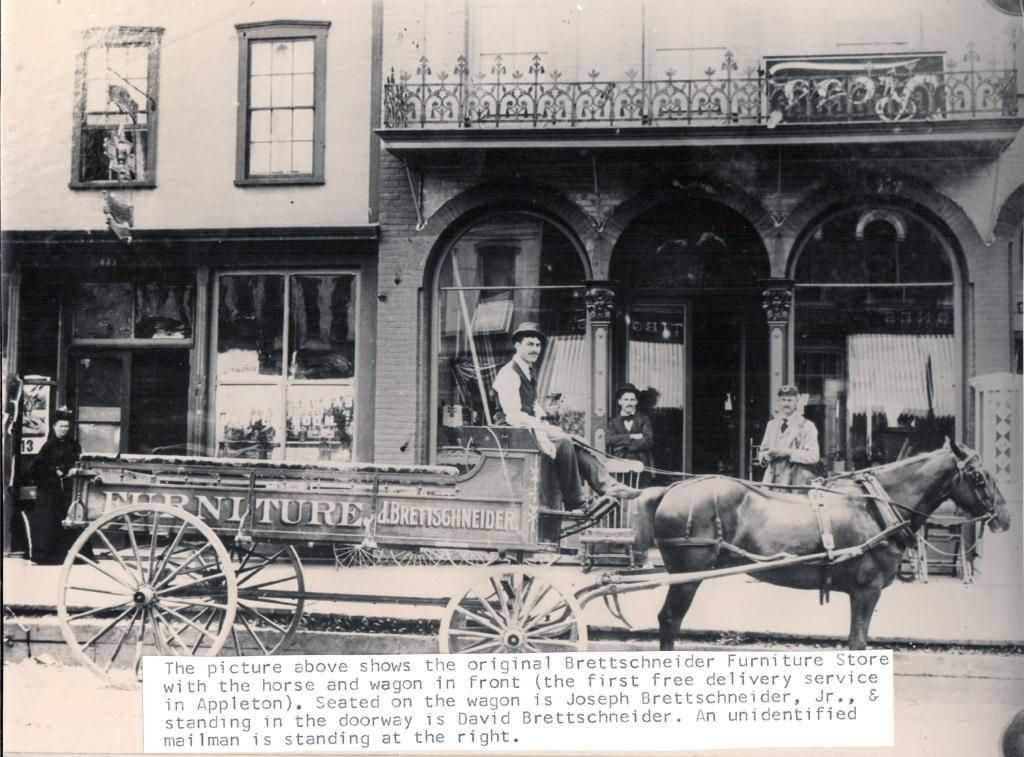 Horse-drawn wagon on a street in front of storefronts and a balcony, with people standing nearby.