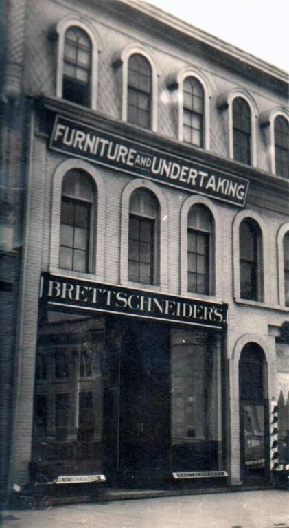 Black-and-white photo of a storefront with signs reading “Furniture Undertaking” and “Brett Schneider”
