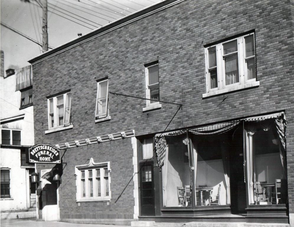 Black-and-white street view of a brick storefront with large display windows and a hanging sign.