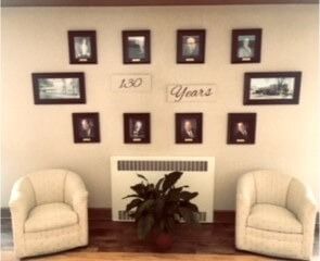 Lobby wall with framed photos, “150 Years” sign, and two beige chairs beside a potted plant
