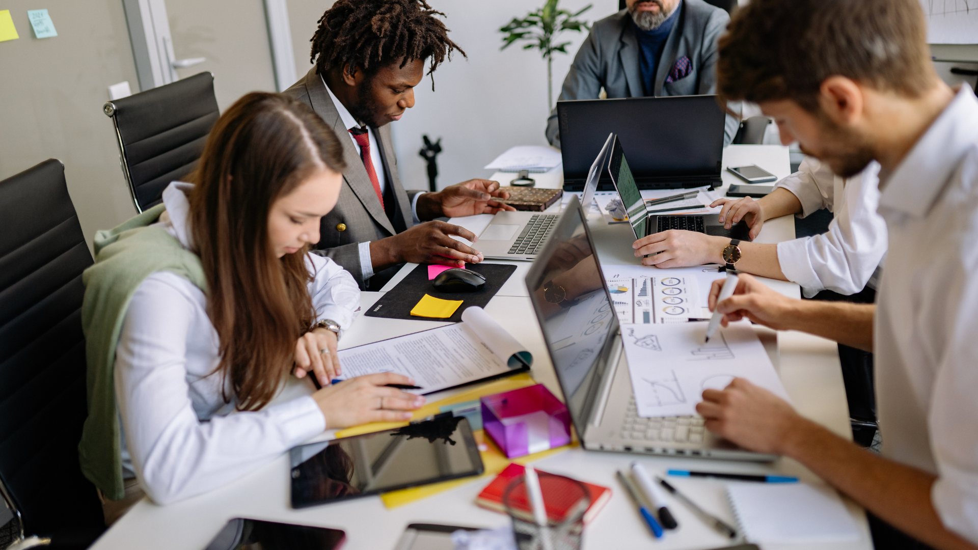 People working together around a table with laptops, papers, and notebooks in an office meeting.