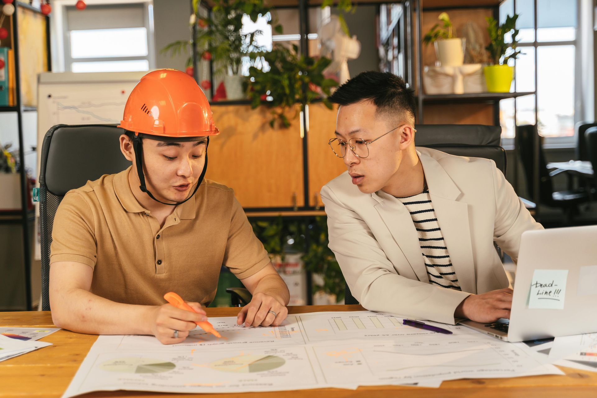 Two coworkers reviewing plans at a desk, one in an orange hard hat, in an office setting