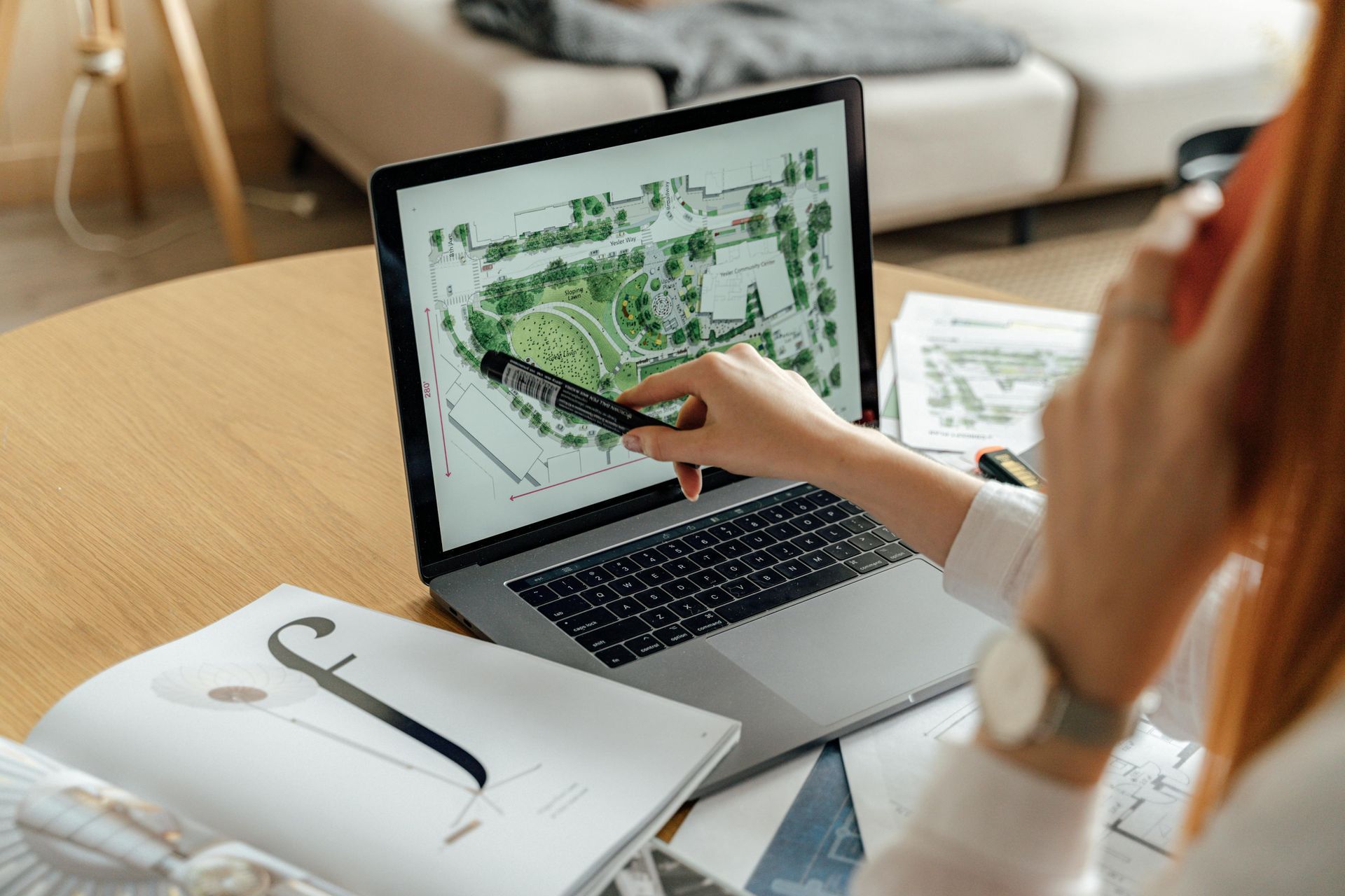Person pointing at a laptop displaying a green design mockup on a desk with sketchbook and tablet