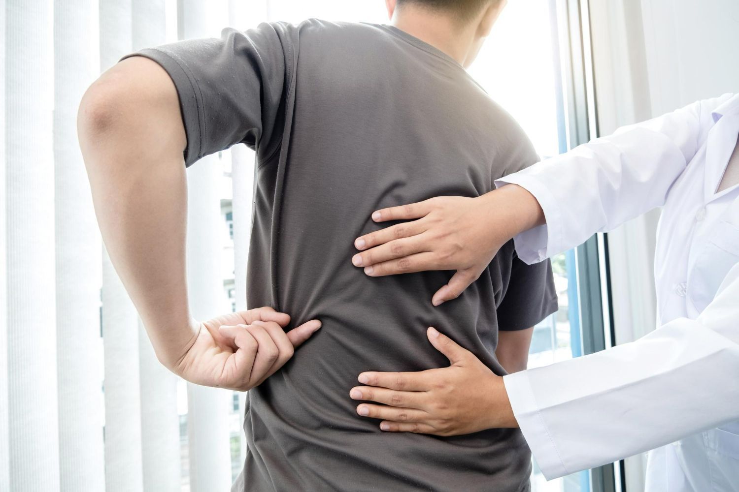 A healthcare professional in a white coat palpates the lower back of a patient wearing a gray shirt.
