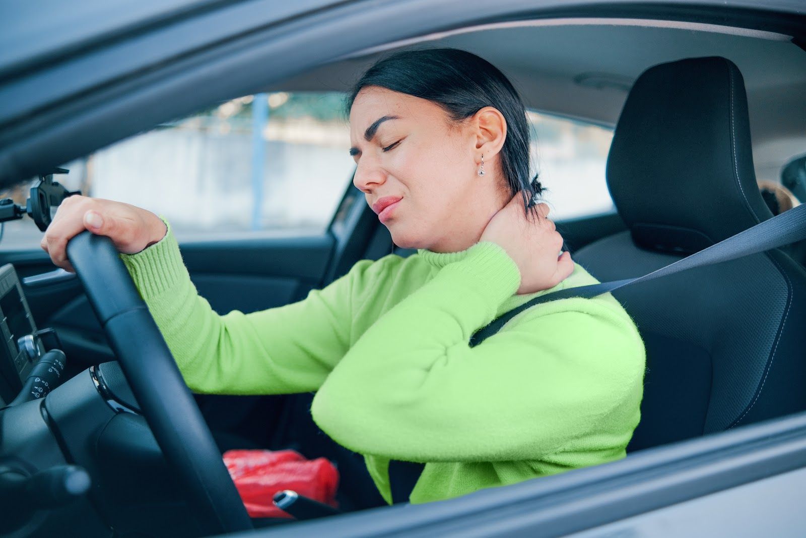Woman in a green sweater rubbing her neck while sitting in a car seat, appearing uncomfortable.