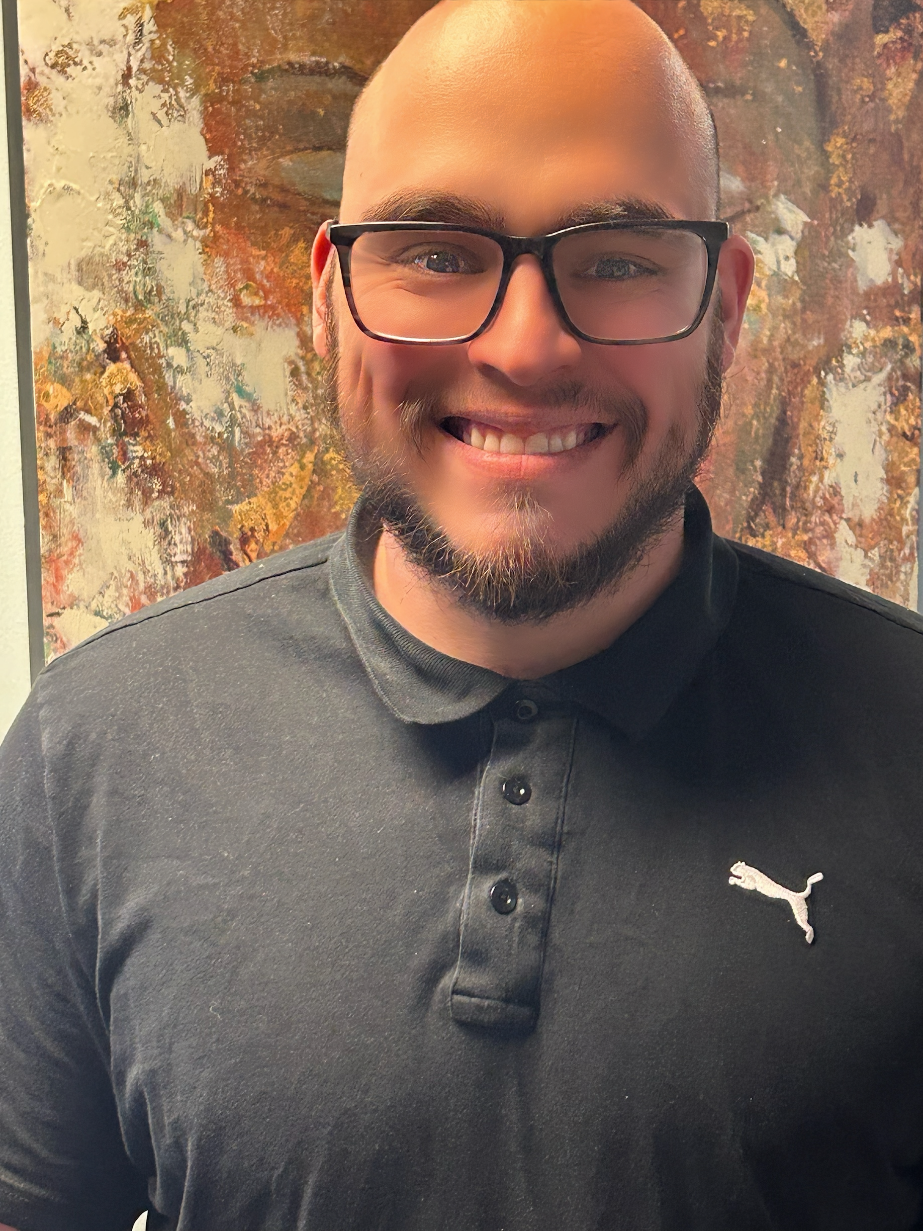 Smiling person with glasses and a dark gray Puma shirt standing indoors against an abstract wall.