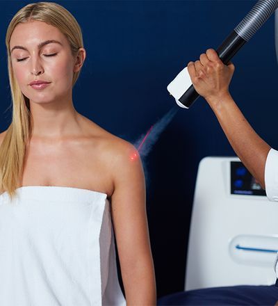 Woman receiving laser treatment on her shoulder in a clinic, with red light targeting a small spot on her skin