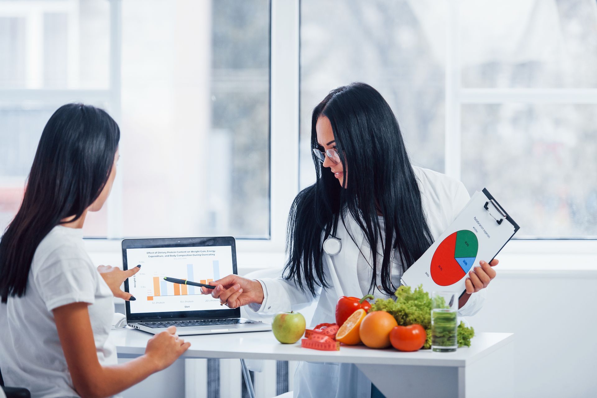 Two people reviewing charts at a table with fruit and a laptop in a bright office