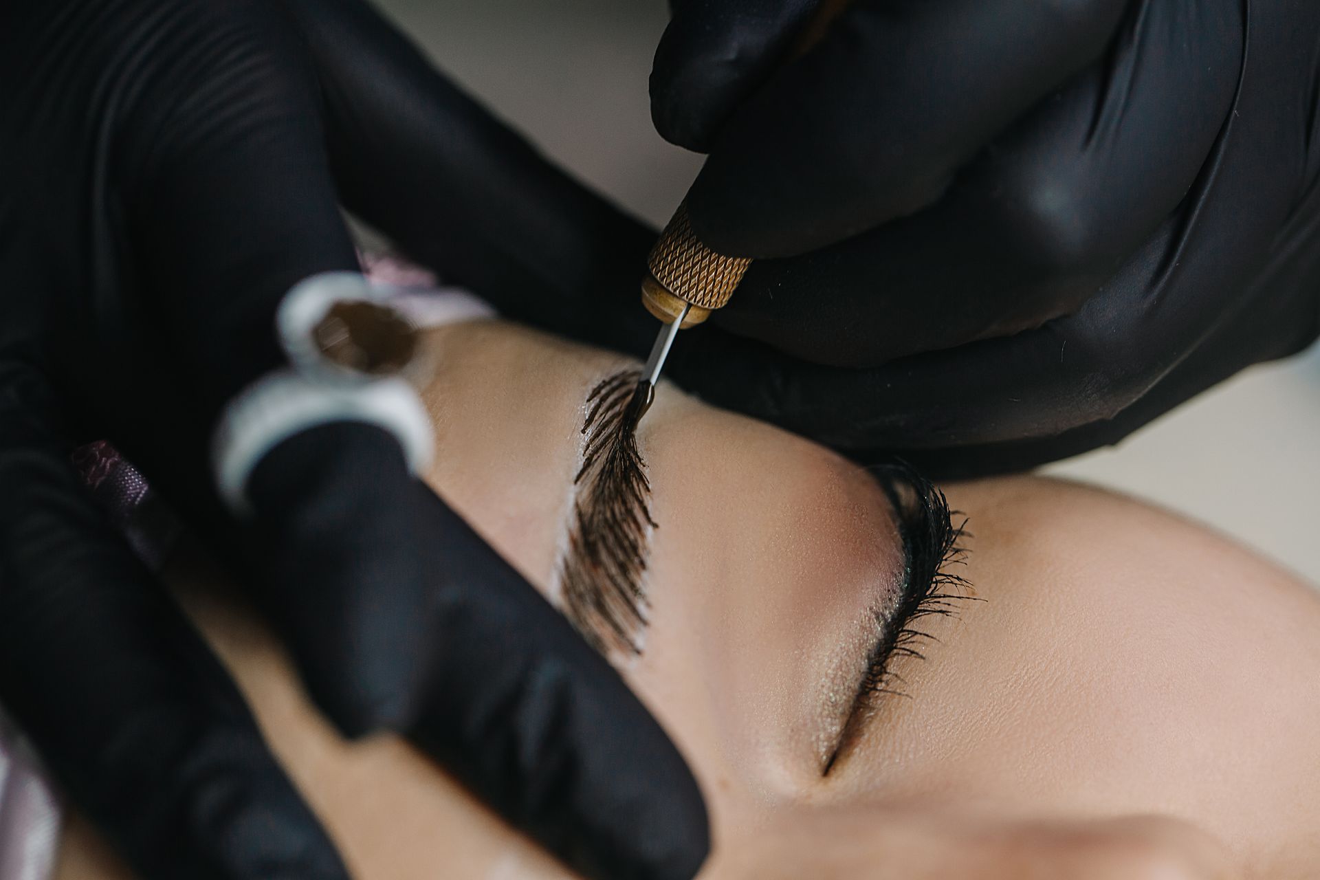 Black-gloved hands applying eyebrow pigment with a brush to a closed eye during cosmetic treatment
