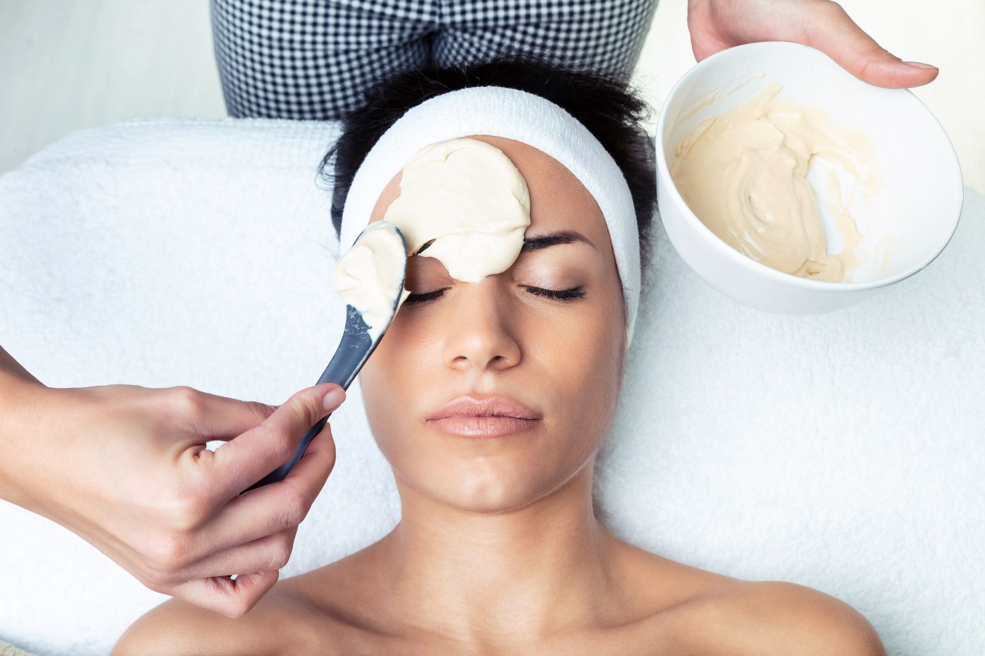 Spa treatment: woman receiving a cream facial mask applied with a brush while another bowl of cream is held nearby