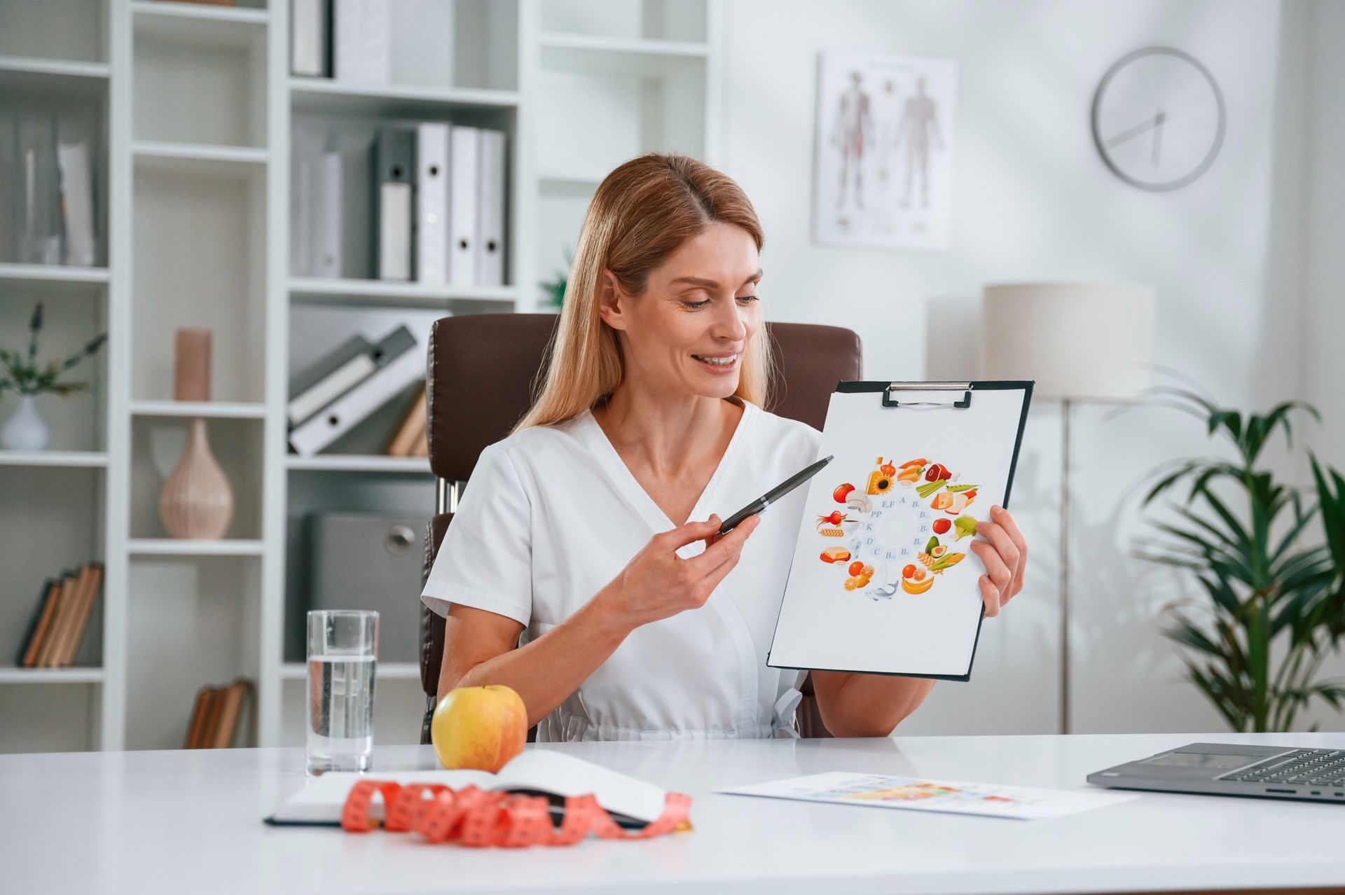 Woman in a white coat holding a clipboard with a food plate diagram in a bright office
