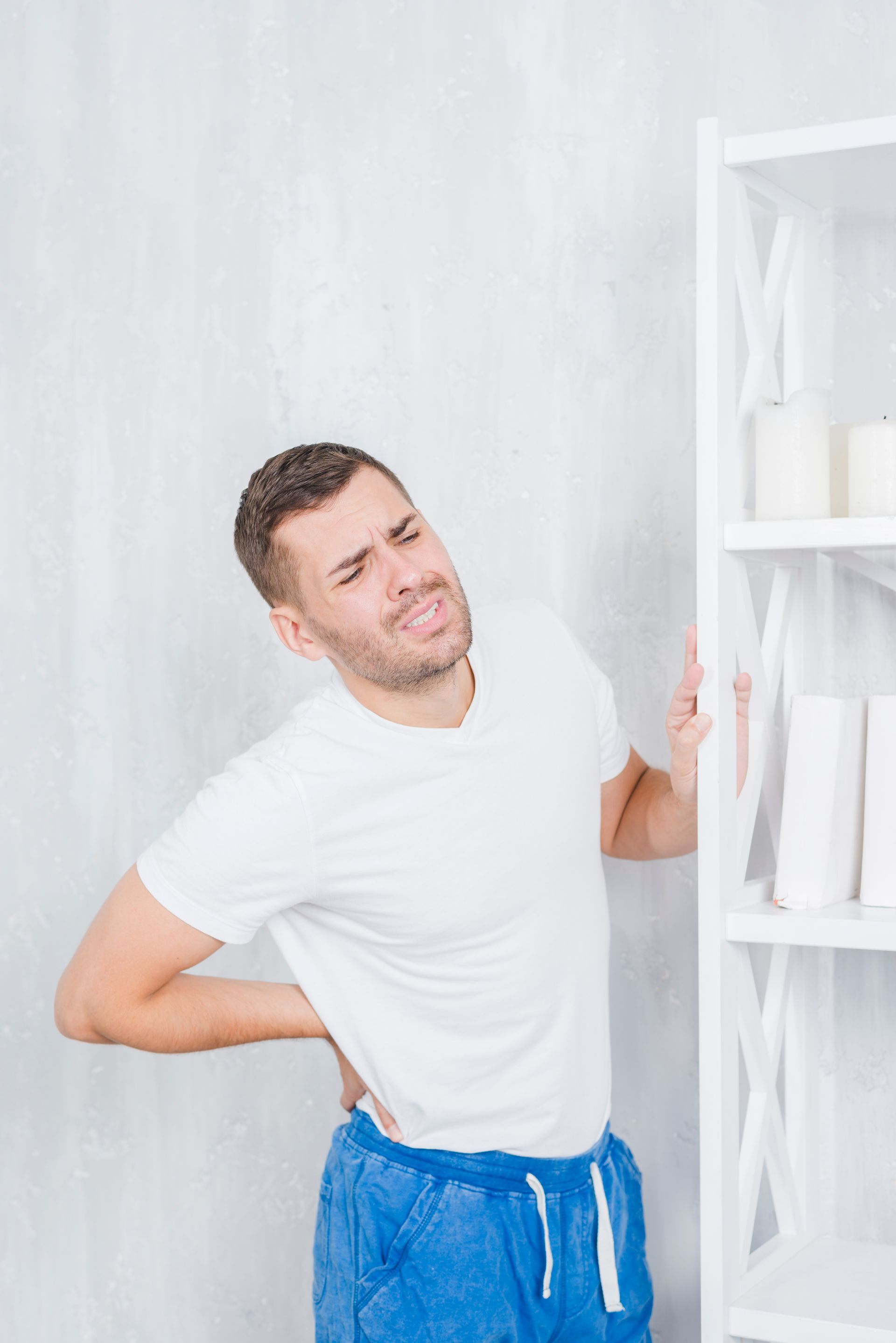 A man is holding his back in pain while standing next to a bookshelf.