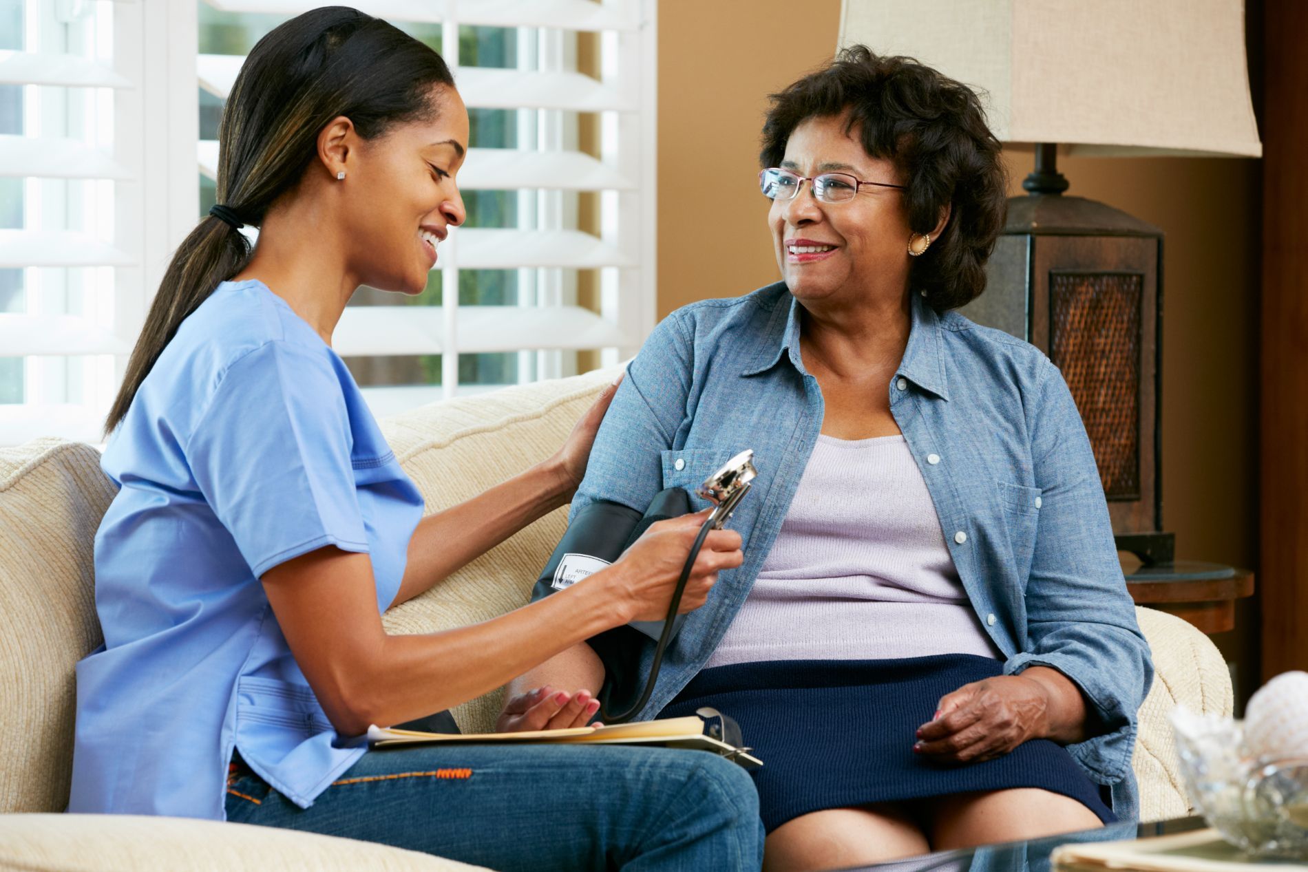 A caregiver in pink scrubs serves breakfast to an elderly woman on a couch; both are smiling