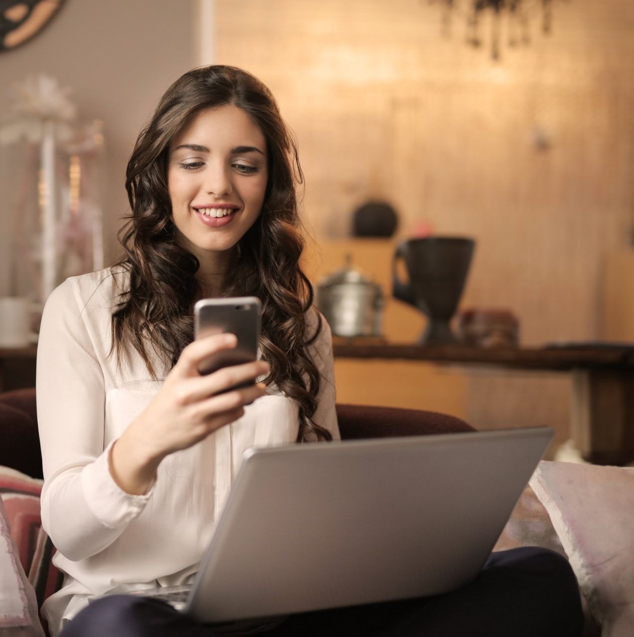 A woman is sitting on a couch using a laptop and a cell phone