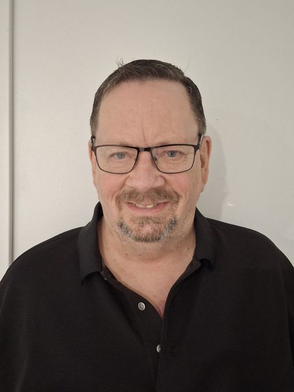 Man wearing glasses and a black polo shirt, smiling against a plain light wall.