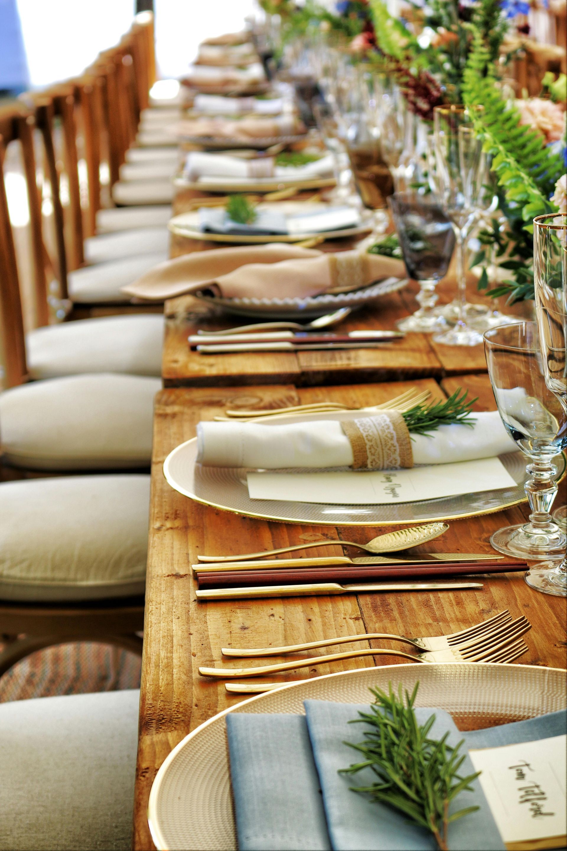 Long wooden banquet table set with woven placemats, white plates, glassware, and greenery centerpiece