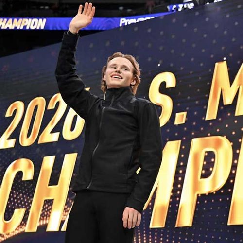Smiling athlete in black jacket waving on stage with 2020 championship sign