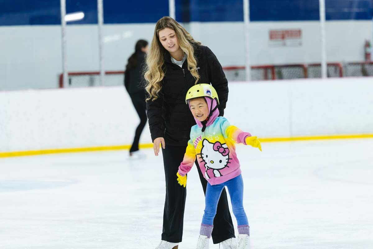 Woman assisting child on ice rink, both wearing skates and helmets