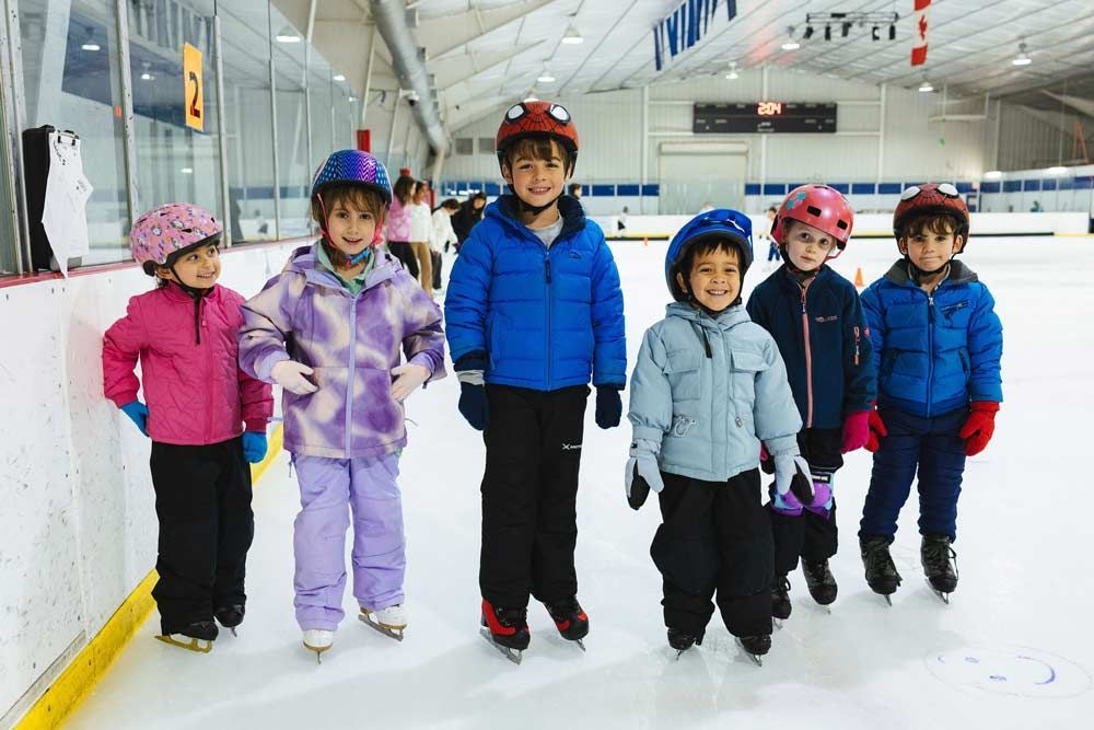 Six children in winter gear standing on an indoor ice rink, holding hands and smiling