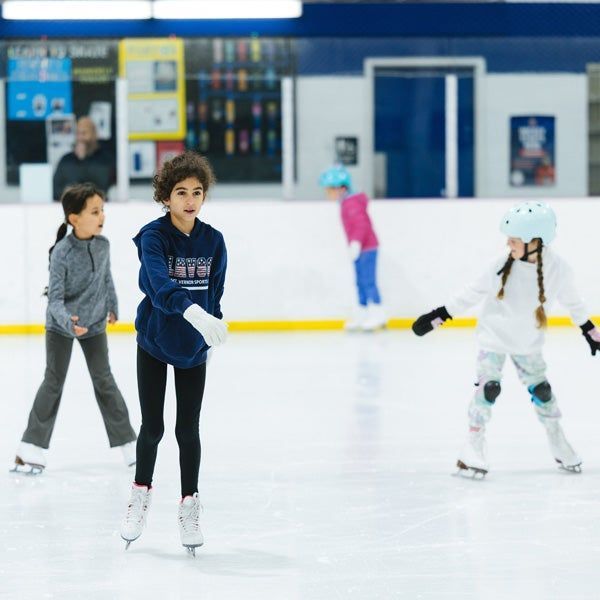 Children ice skating at an indoor rink, with one in a blue sweater in the foreground