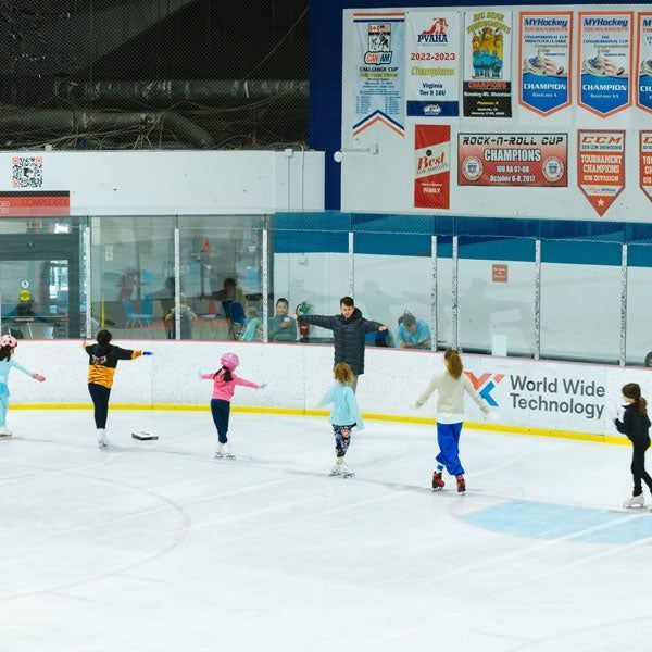 Children holding hands skate in a line on an indoor ice rink with a coach nearby.
