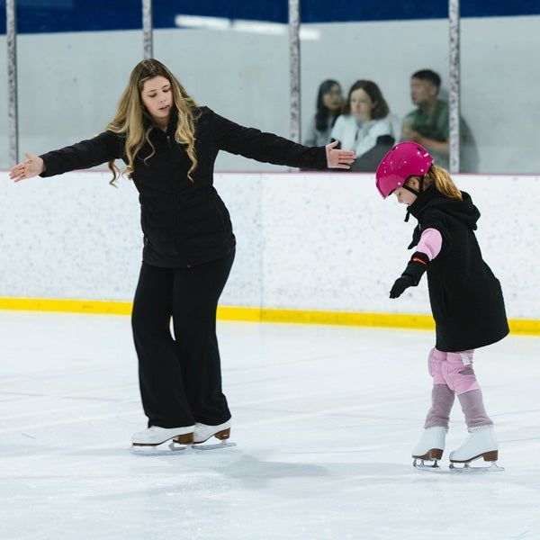 Woman and child ice skating together indoors, holding arms out for balance.